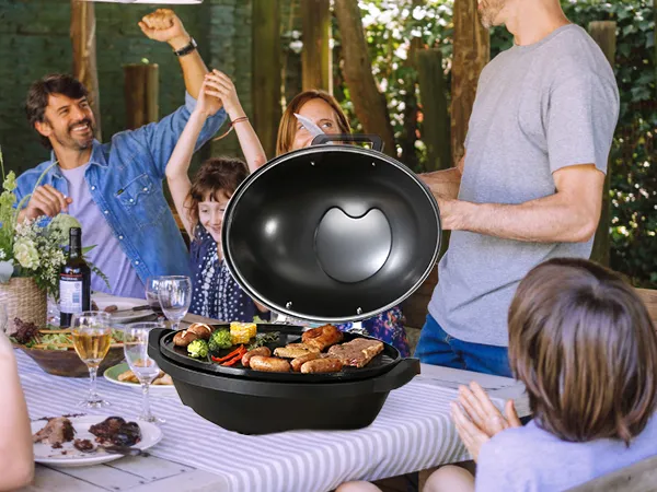 Reunión familiar alrededor de una mesa con una parrilla eléctrica VEVOR, llena de carnes y verduras a la parrilla.