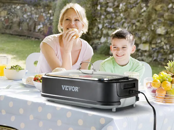 Familia disfrutando de una barbacoa al aire libre con una parrilla interior sin humo VEVOR sobre una mesa, con un frutero colorido cerca.