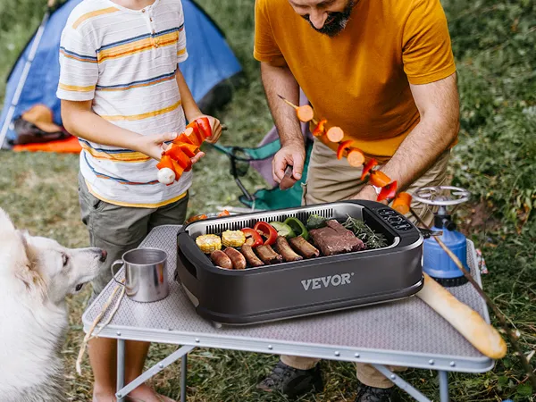 Escena de campamento con parrilla interior sin humo VEVOR sobre una mesa, brochetas de comida, un perro y una fogata.