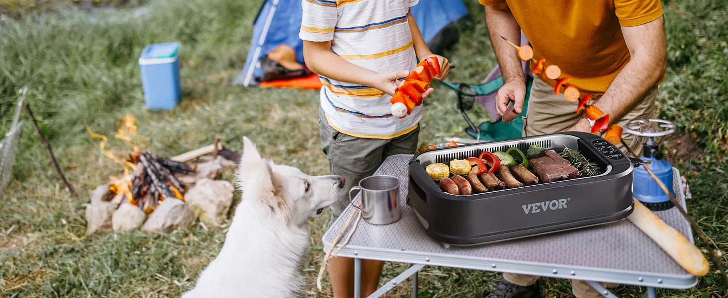 Escena de campamento con parrilla interior sin humo VEVOR sobre una mesa, brochetas de comida, un perro y una fogata.