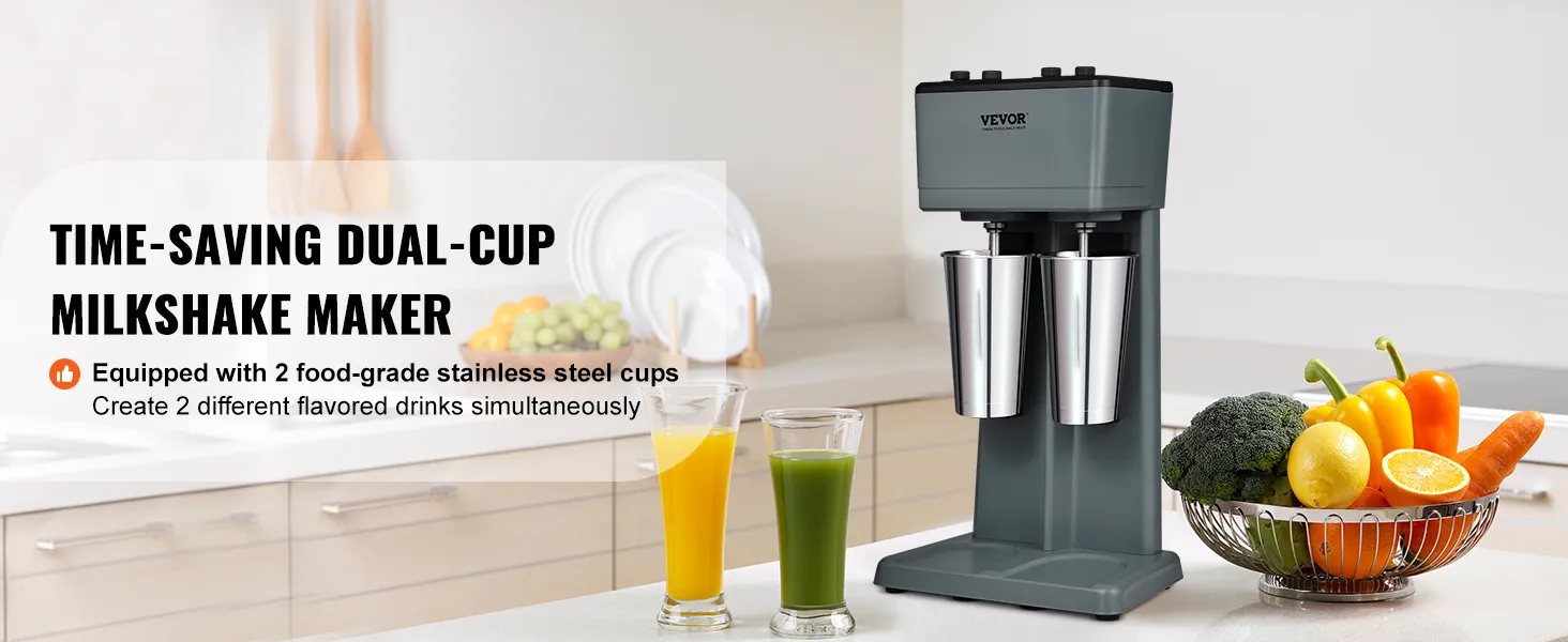 Máquina para hacer batidos VEVOR en una cocina moderna con dos tazas de acero inoxidable y verduras frescas cerca.