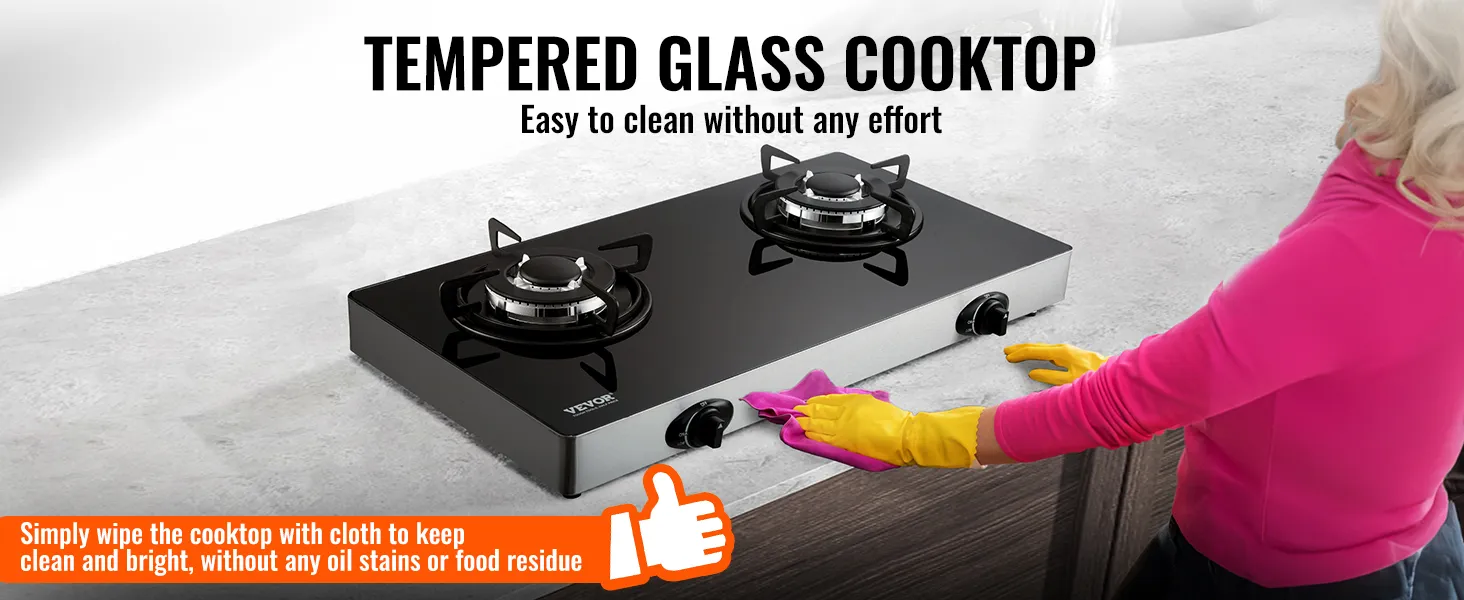 Cocina a gas VEVOR con vidrio templado, limpiada por una persona con sudadera rosa y guantes amarillos.