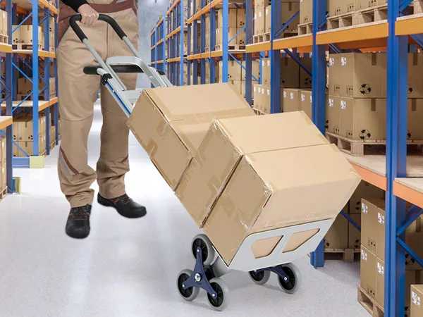 Un trabajador del almacén utiliza un carrito sube escaleras VEVOR para mover cajas a través de estantes de metal llenos de paquetes.